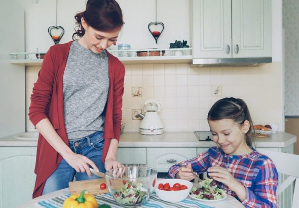 Happy mother and cheerful daughter cook salad together in the kitchen at home cutting vegetables and talking. Family, cooking, and people concept